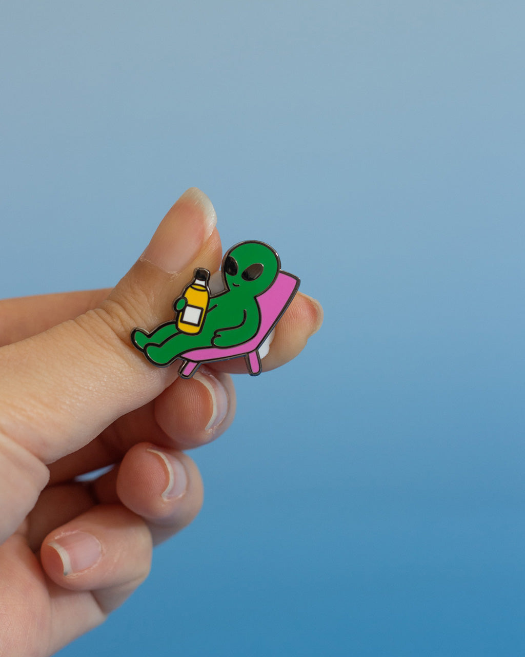 Hand holding an enamel pin that has a design of a green alien against a blue background