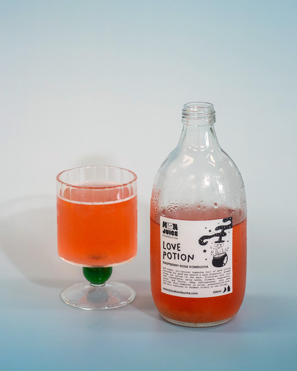 A half full bottle of kombucha labelled "Love Potion", and a glass of kombucha on a blue gradient background
