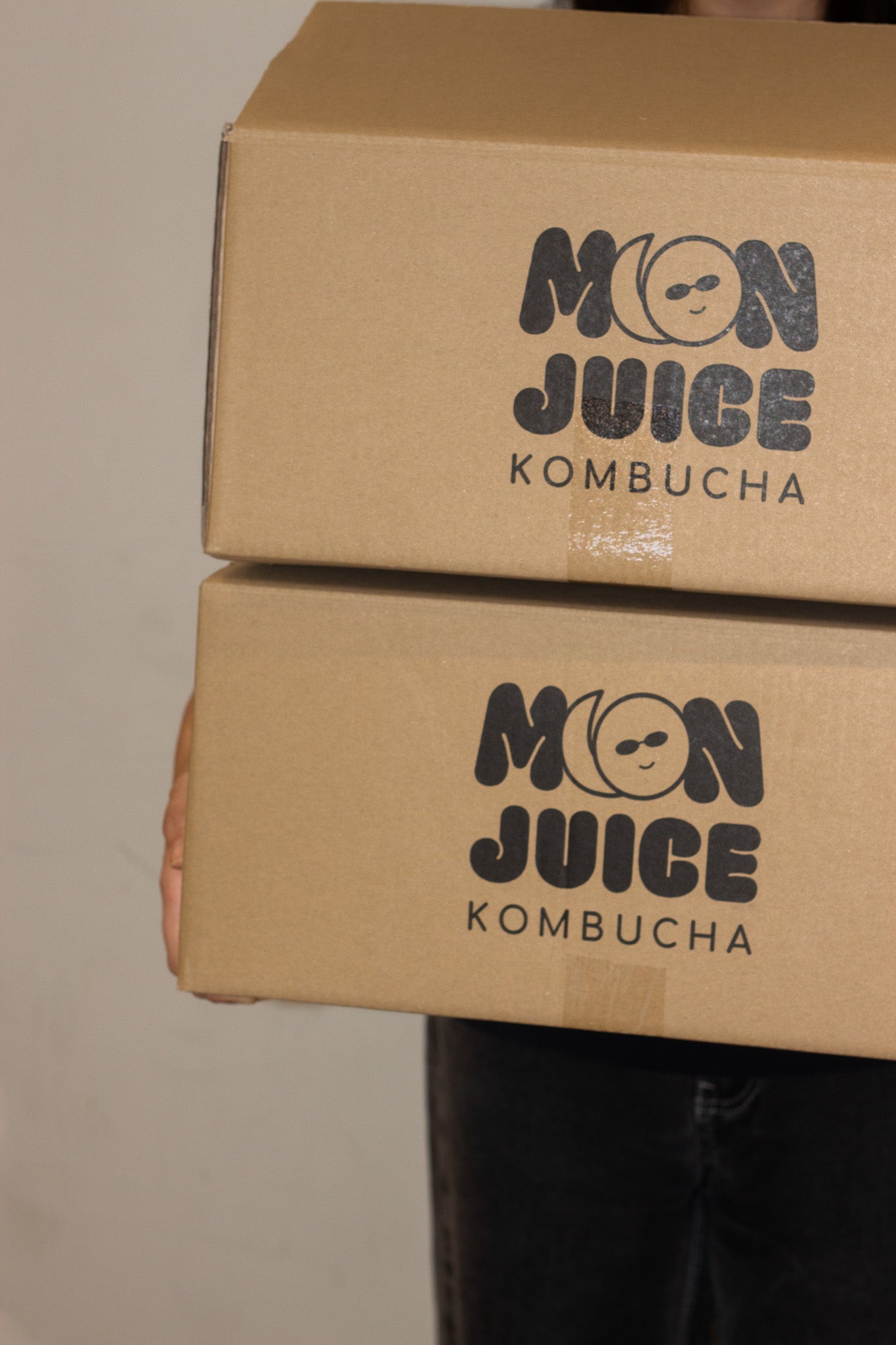 A person carrying 2 cardboard boxes with 'Moon Juice Kombucha' branding on a neutral background.