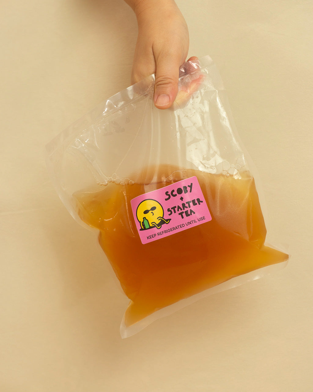 A person holding a SCOBY + Starter tea packet with a pink label against a beige background