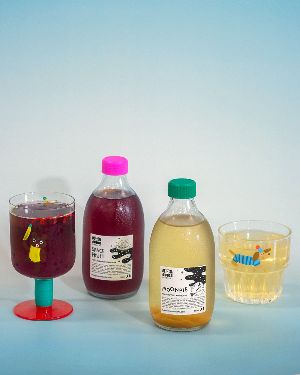 Two bottles of kombucha with a pink & a green cap, and two glasses filled with a purple kombucha, and a yellow kombucha on a light blue background.