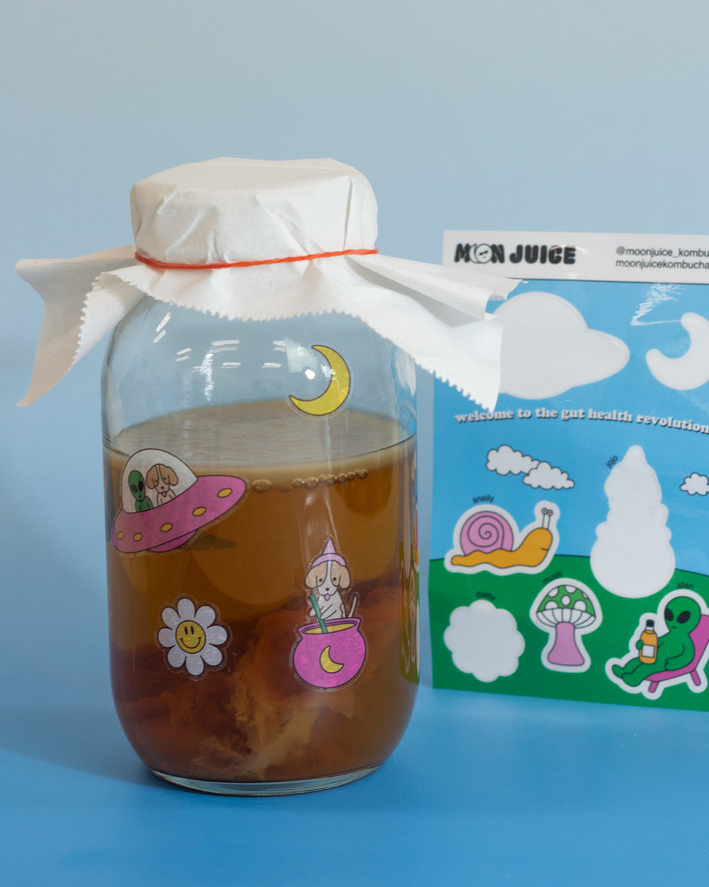 A jar of kombucha with 4 Moon Juice Kombucha stickers on it, next to a half-empty sticker sheet