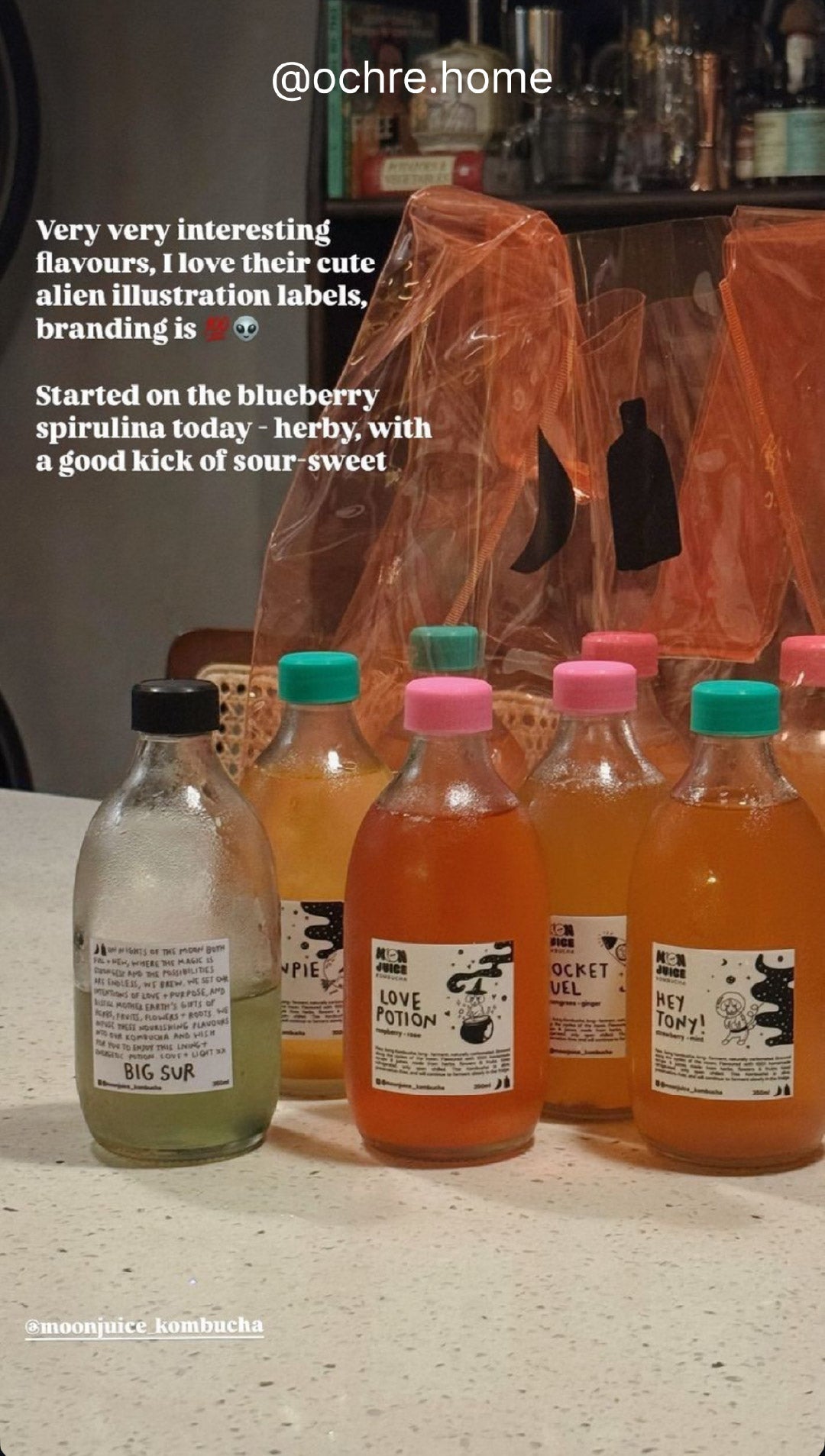 Lifestyle shot of 7 full kombucha bottles and 1 half-full Big Sur bottle on a table, with a orange Moon Juice jellybag in the background. Taken by @ochre.home