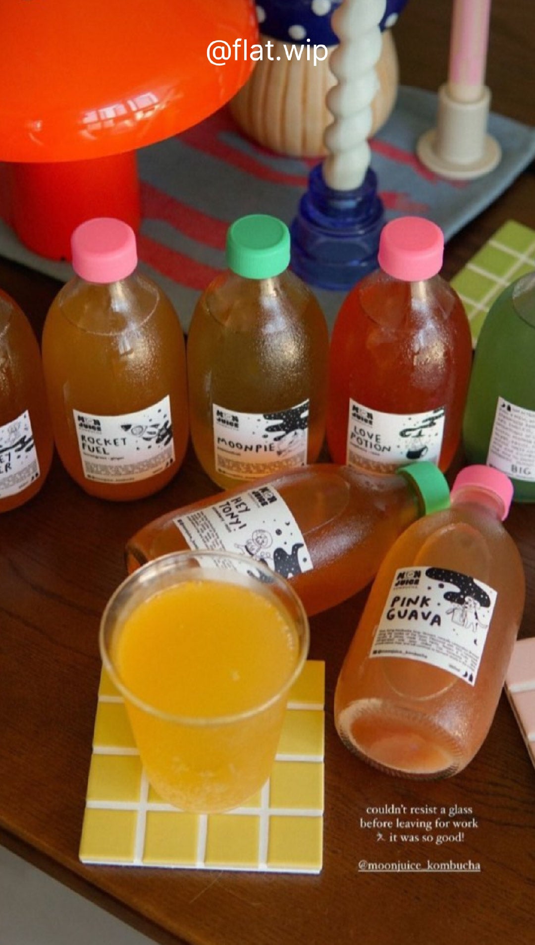 Aesthetic lifestyle shot of 7 kombucha bottles and a cup on a table with coasters, a lamp, some candles. Taken by @flat.wip