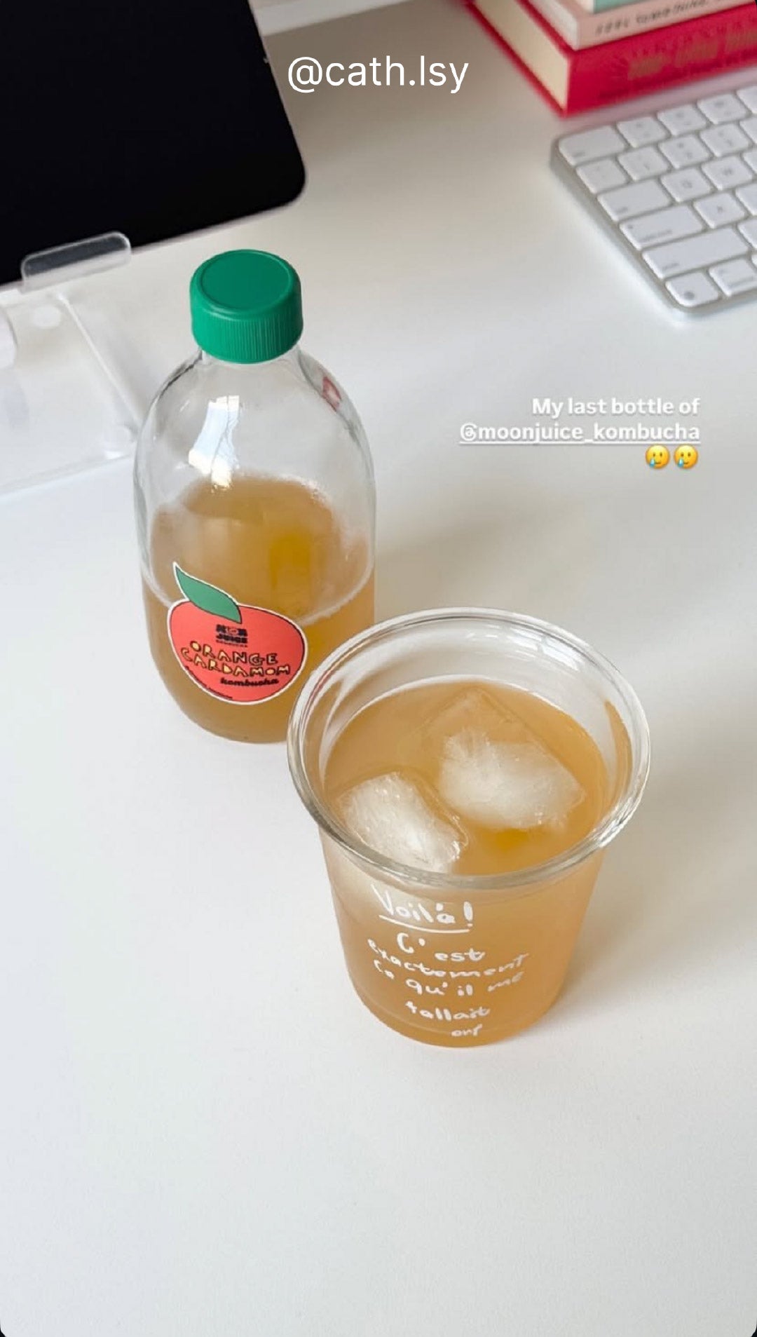 A half-full bottle of special flavour Orange Cardamom next to a cup of kombucha with ice, placed on a workdesk. Taken by @cath.lsy