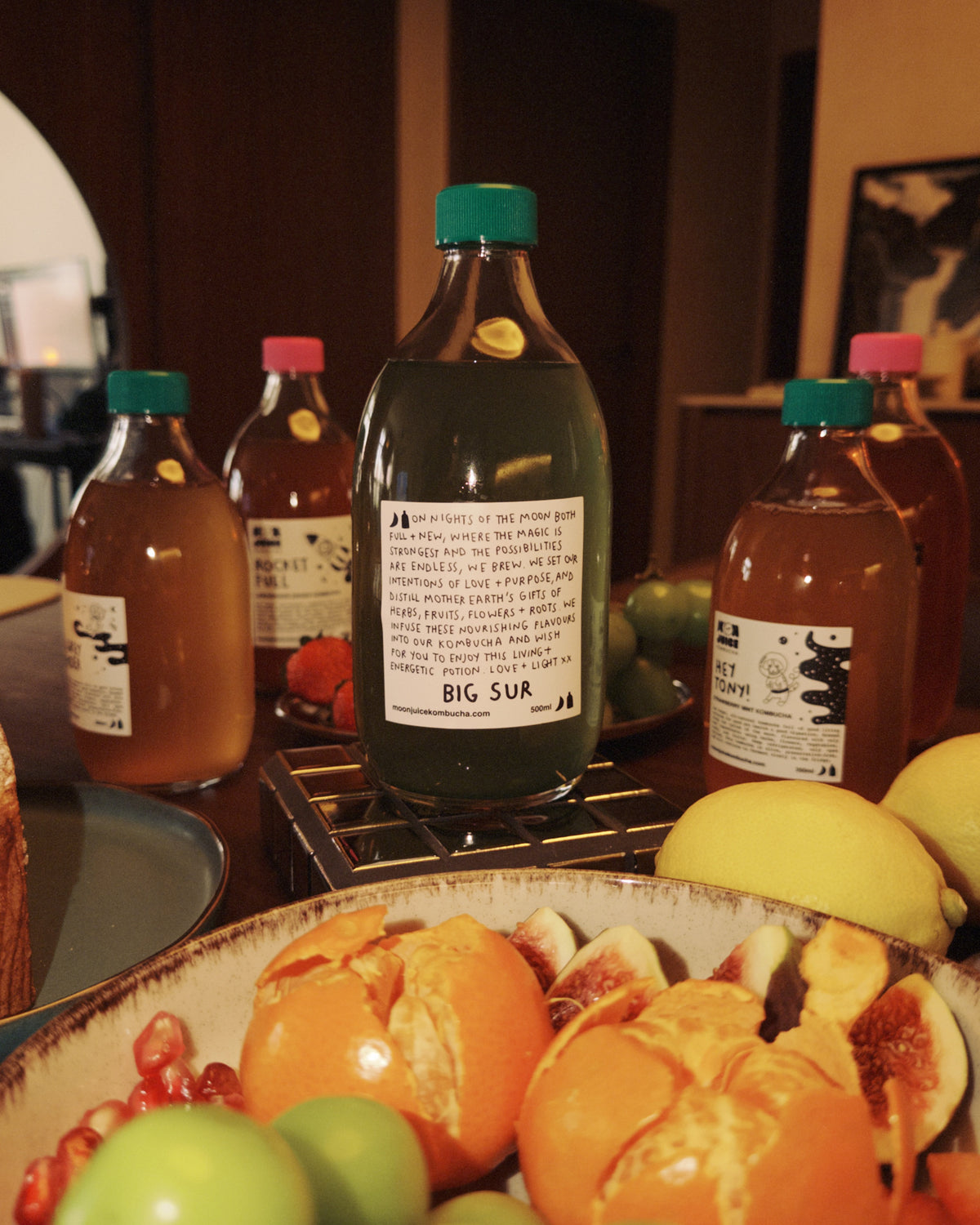 A bottle of Big Sur kombucha surrounded by other kombucha bottles and a platter of fruits on a table.