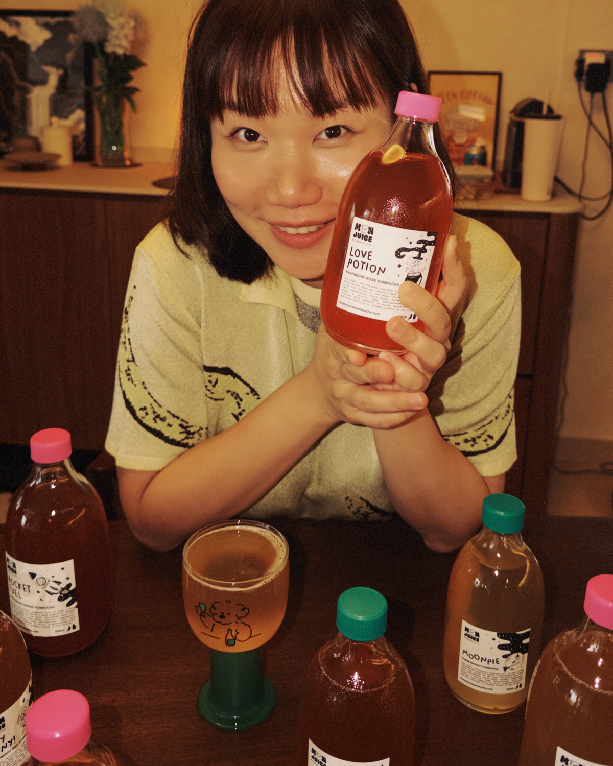A woman smiling and holding up a bottle of Love Potion bottles. Several similar kombucha bottles are arranged on a wooden table in front of her.