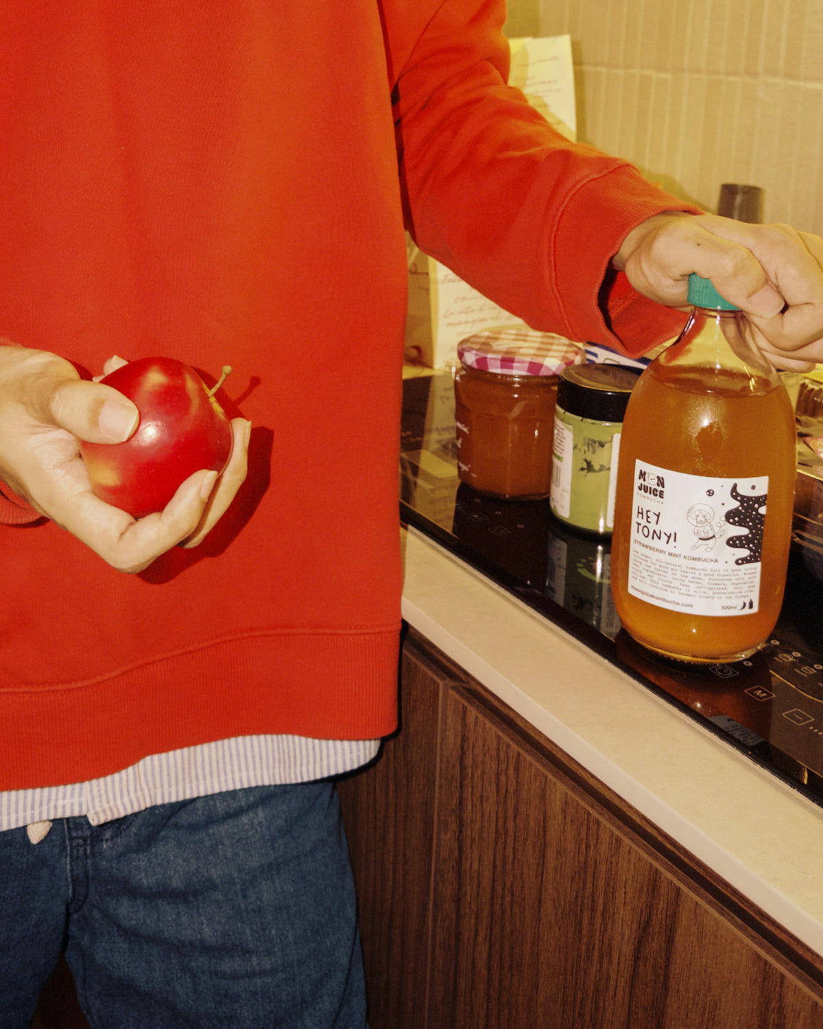 A person in a red sweatshirt holds a red apple in one hand and a large bottle of Hey Tony! kombucha standing at a kitchen counter with jars in the background.