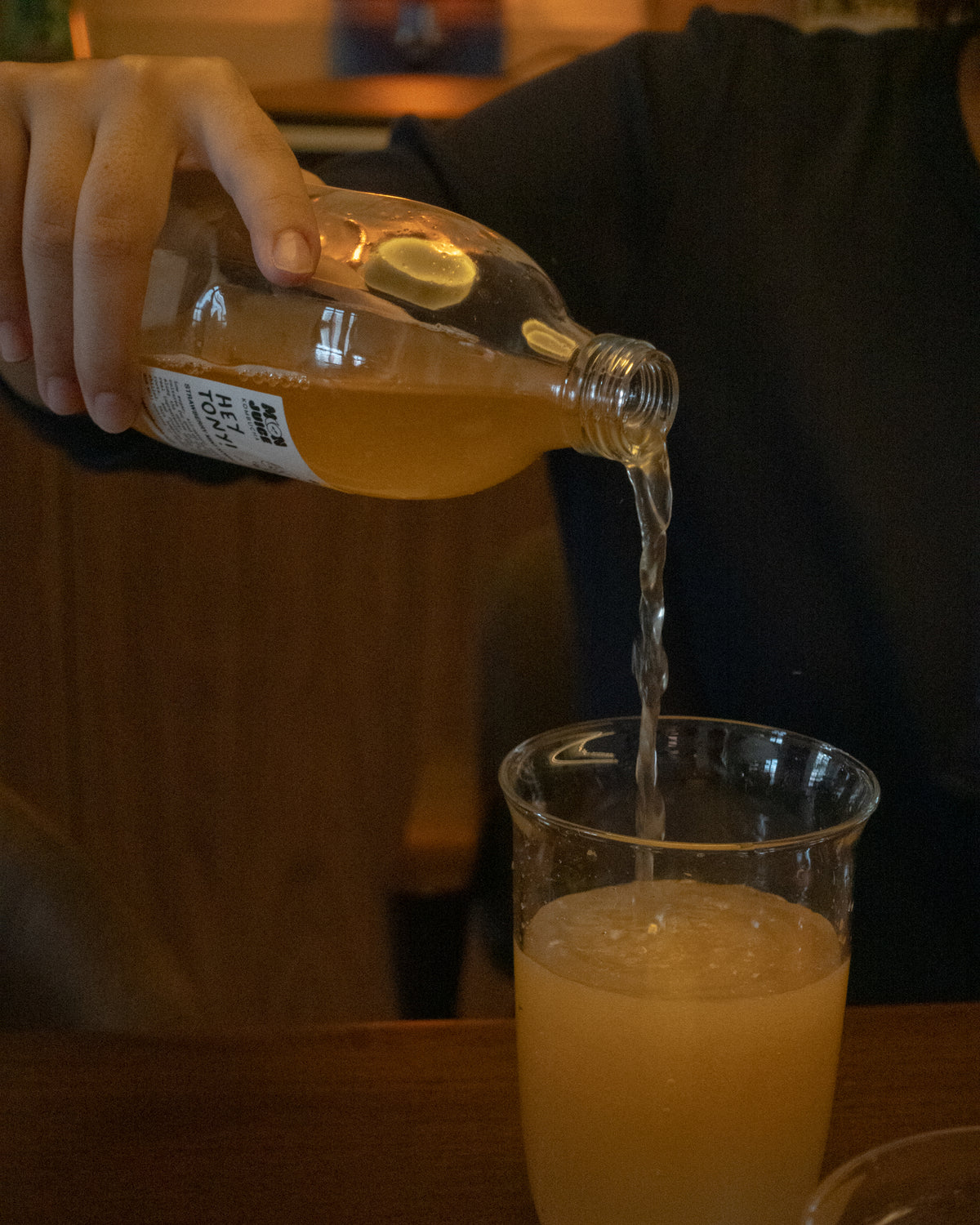 A person pouring a bottle of Hey Tony! kombucha into a clear glass on a wooden table in a dim indoor setting.