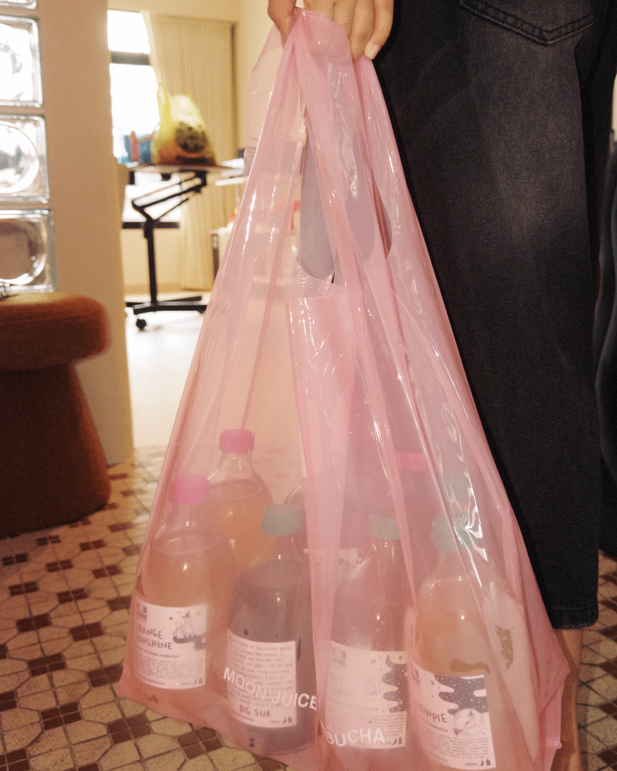 A person holding a translucent pink jellybag filled with 8 kombucha drinks in an indoor room with patterned tile flooring and furniture in the background.