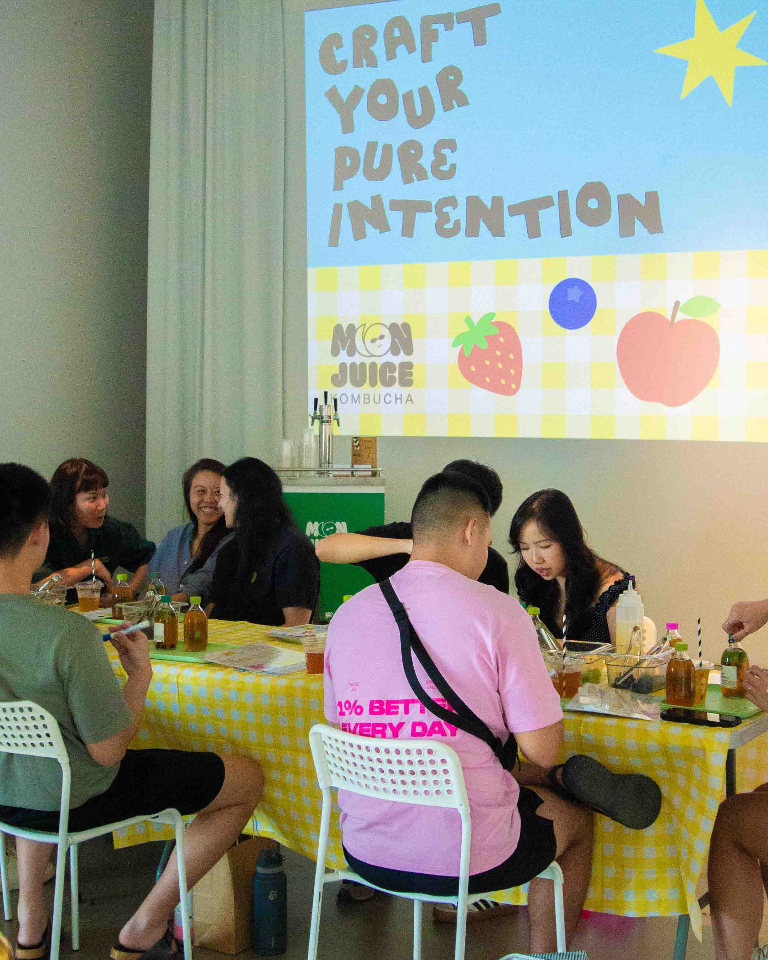 Group of people sitting at a table with a projector screen displaying 'Craft Your Pure Intention' and 'Moon Juice Kombucha' branding.