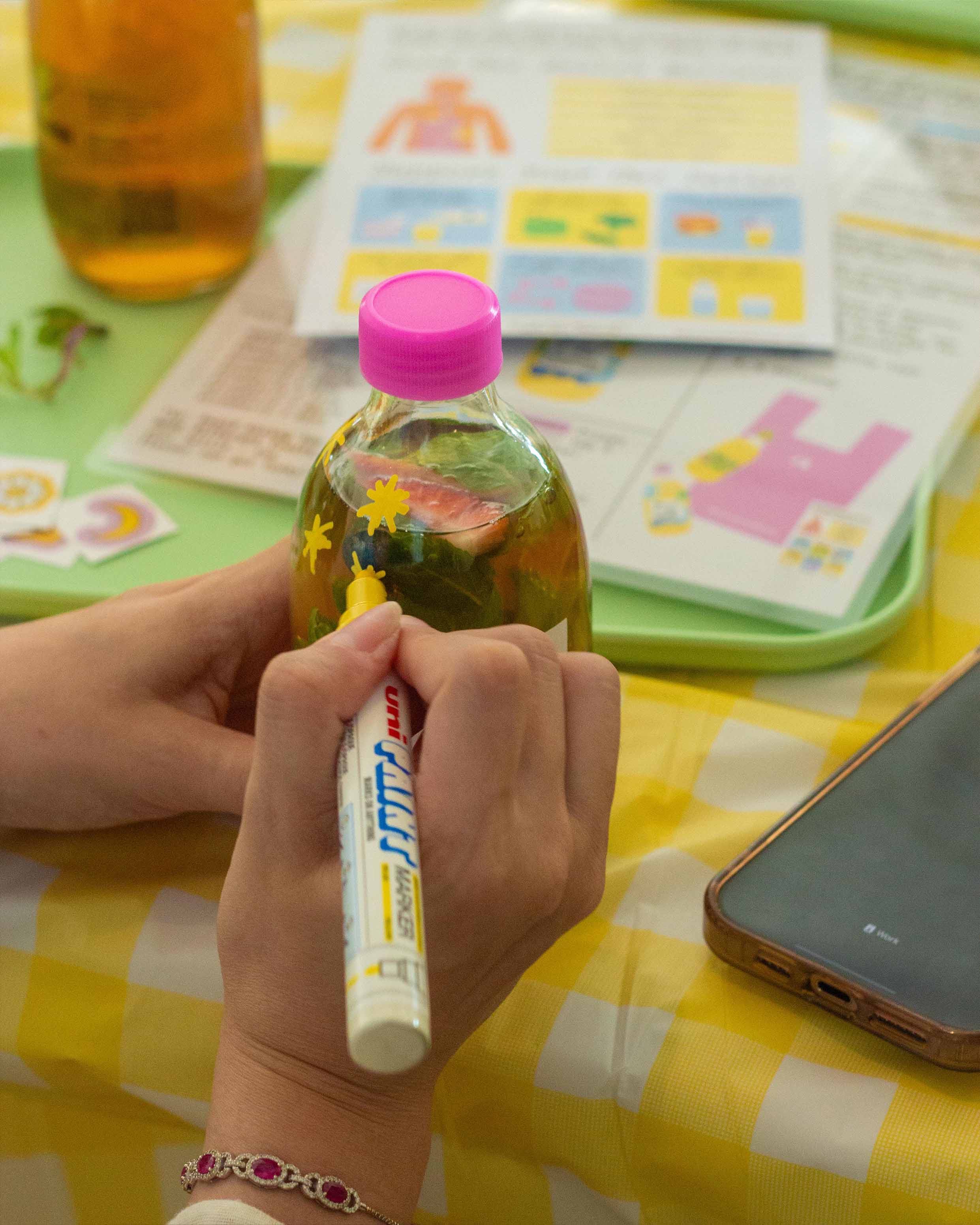 Person decorating a bottle of kombucha using a paint marker, against a yellow checkered tablecloth.