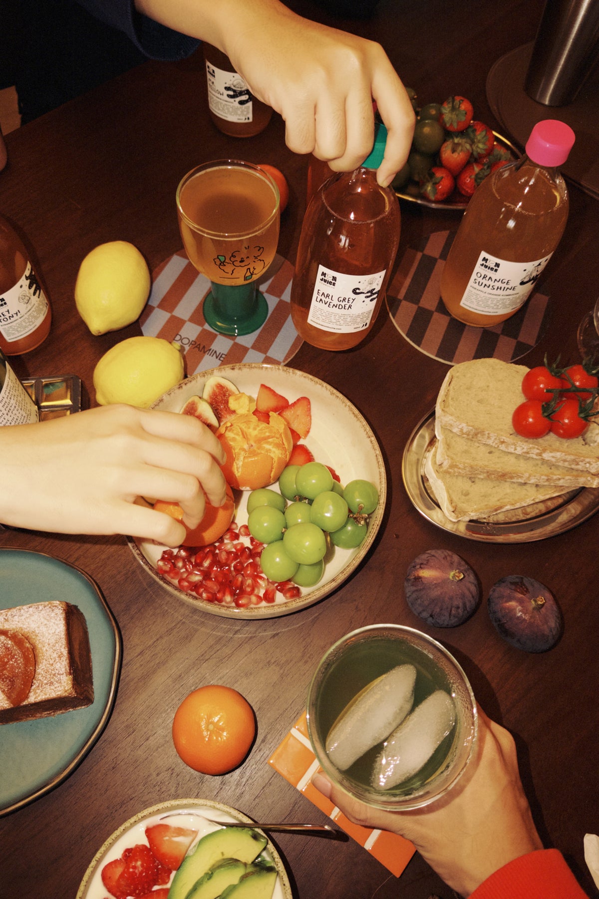 A cozy tabletop scene with multiple hands serving and drinking from kombucha bottles, alongside plates of sliced fruit, grapes, bread, and a drink with ice on a dark wooden table.