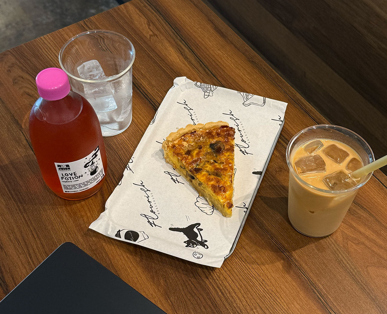 A bottle of Love Potion kombucha, with a cup of ice, a slice of quiche, and a cup of coffee next to it.