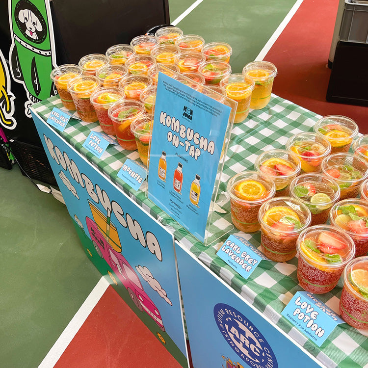 Dual Kombucha Cart set up outdoors with a table displaying rows of kombucha in cups. Featuring foam board cut outs for branding & decor!