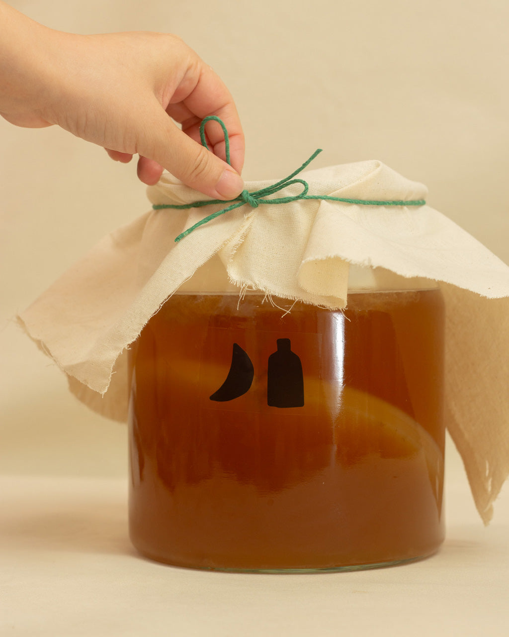 Jar of kombucha with a scoby inside it on a beige background. A hand ties a green string around the cloth cover of the jar.