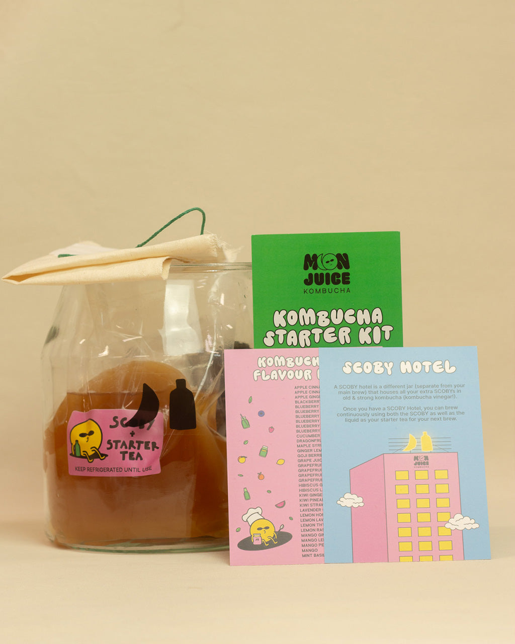 Kombucha starter kit on a beige background - includes a glass jar, tea bags, SCOBY + starter tea, tea leaves + sugar, linen cloth + string, and 3 instruction sheets.