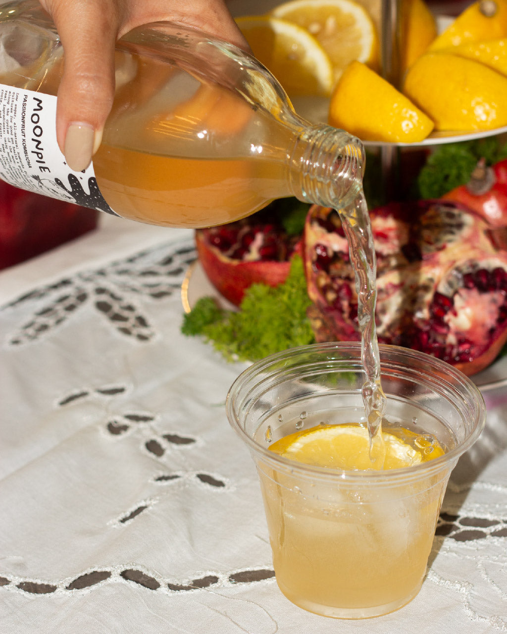 Hand pouring kombucha from a bottle labeled 'Moonpie' into a clear cup with lemons and pomegranates in the background.