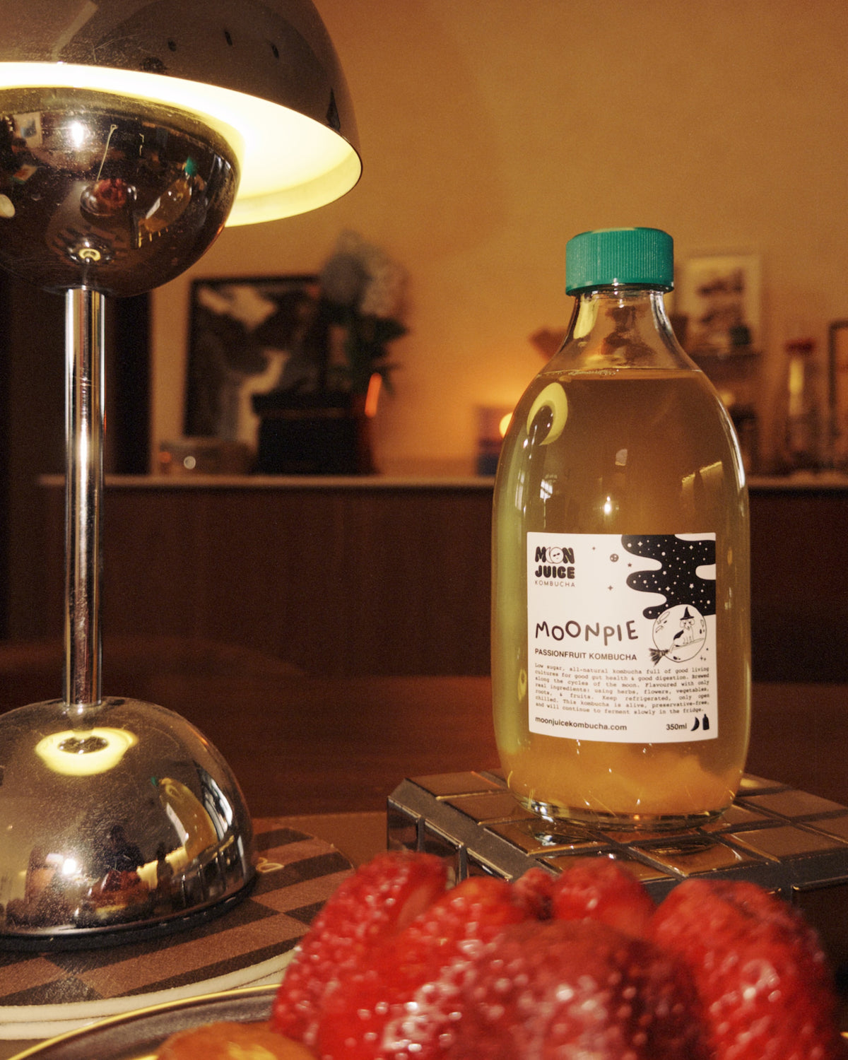 A bottle of Moonpie kombucha on a wooden table next to a lamp, and a strawberry danish