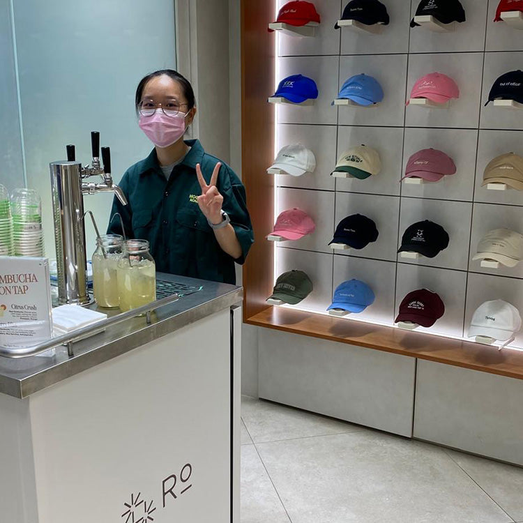 A kombucha cart with a staff holding a peace sign next to a display of colorful caps on a wall-mounted shelf