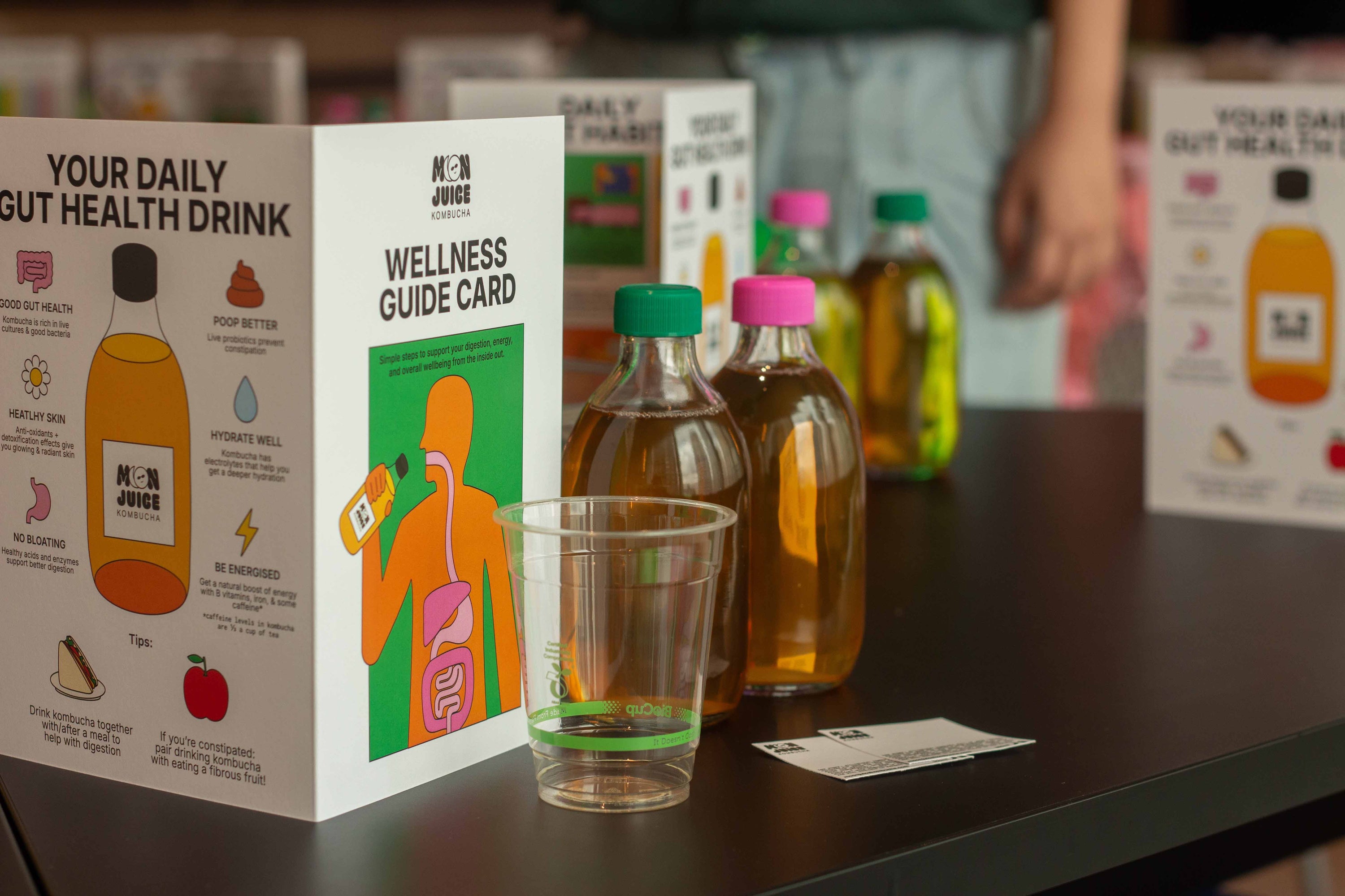 A table with our kombucha wellness workshop set-up: a wellness guide card, a cup, 2 blank stickers, and 2 bottles of kombucha.