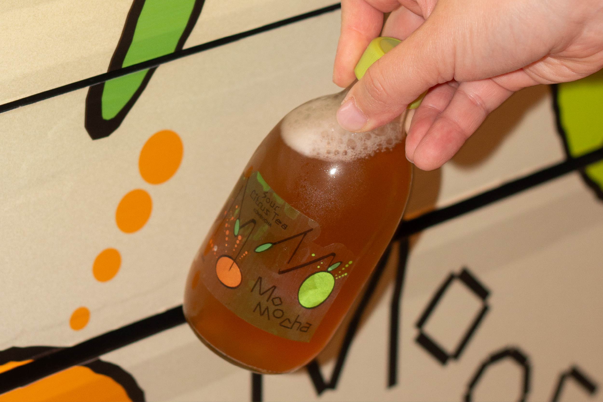 Hand holding a bottle of 'Momocha' kombucha against a patterned background of a vending machine
